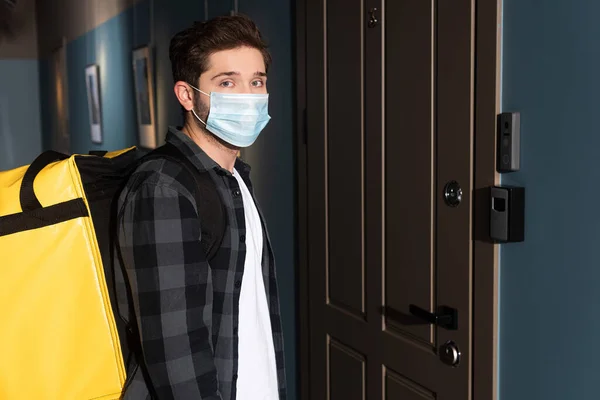 Side view of courier in medical mask with thermo backpack looking at camera near door in entryway — Stock Photo