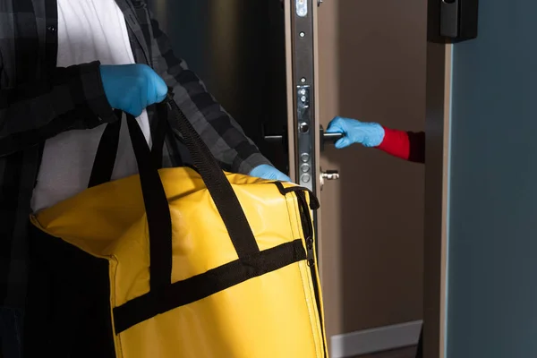 Vista ritagliata dell'uomo di consegna che tiene la borsa termica vicino alla donna in guanto di lattice porta di apertura — Foto stock