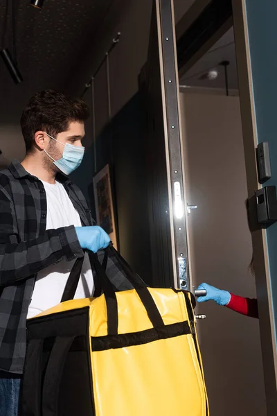 Side view of courier in medical mask holding thermo bag near woman in latex glove opening door — Stock Photo
