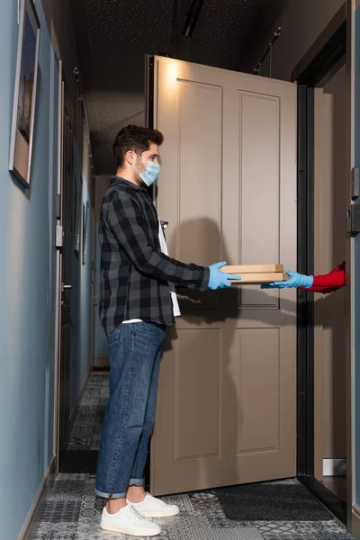 Vista lateral del mensajero en máscara médica dando cajas de pizza a la mujer en guantes de látex cerca de la puerta abierta en la entrada - foto de stock