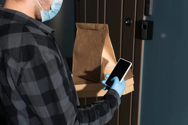 Cropped view of courier in medical mask holding package, pizza boxes and smartphone near door in entryway — Stock Photo