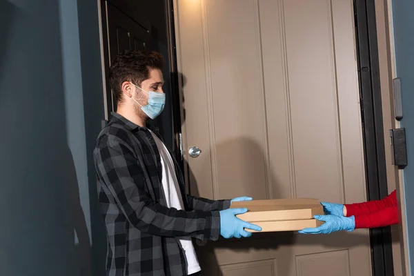 Vista lateral del mensajero en máscara médica dando cajas de pizza a la mujer en guantes de látex cerca de la puerta abierta en la entrada - foto de stock