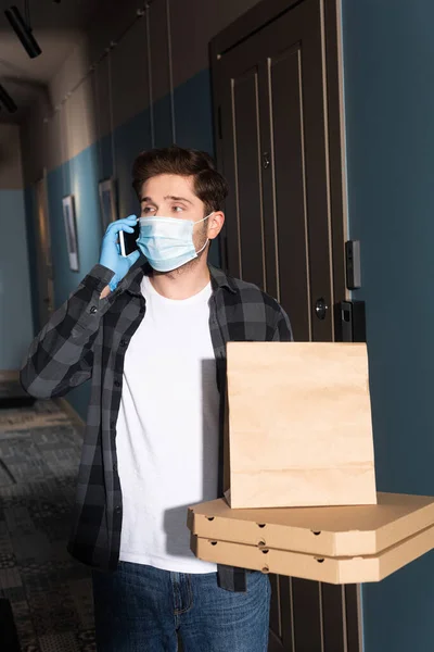 Entrega hombre en máscara médica sosteniendo cajas de pizza y paquete mientras habla en el teléfono inteligente en la entrada - foto de stock