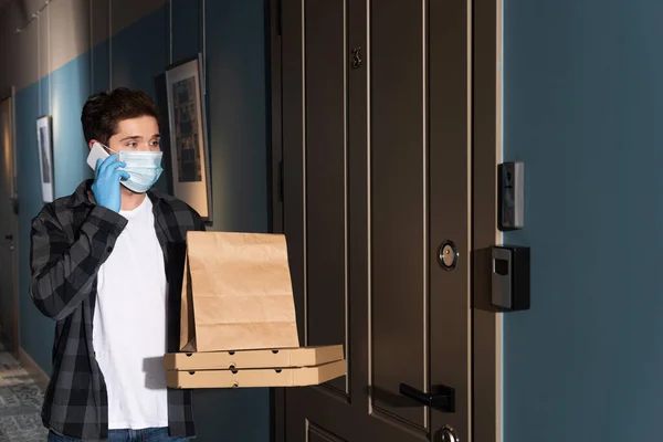 Курьер в медицинской маске разговаривает по смартфону, держа пакет и коробки с пиццей на входе — стоковое фото