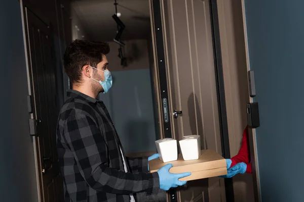 Vista lateral del repartidor hombre en máscara médica dando paquetes y cajas de pizza a la mujer en guantes de látex cerca de la puerta abierta - foto de stock