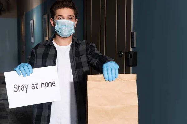 Delivery man in medical mask and latex gloves holding package and card with stay at home lettering on entryway — Stock Photo