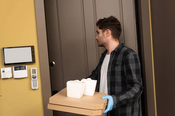 Bonito homem de luva de látex segurando pacotes e caixas de pizza perto da porta aberta — Fotografia de Stock