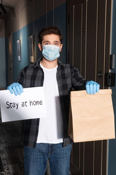 Courier in medical mask and latex gloves holding card with stay at home lettering and package on porch — Stock Photo