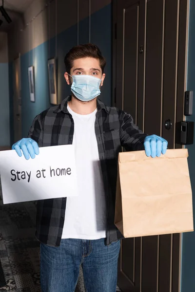 Correio em máscara médica e luvas de látex cartão de segurança com estadia em casa lettering e pacote no alpendre — Fotografia de Stock