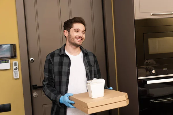 Hombre sonriente con guantes de látex sosteniendo cajas de comida para llevar y pizza en casa - foto de stock