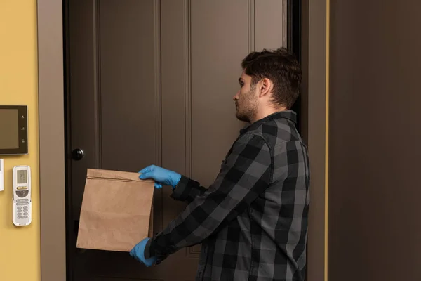 Вид сбоку на красивого мужчину в латексных перчатках, держащего пакет возле открытой двери — стоковое фото