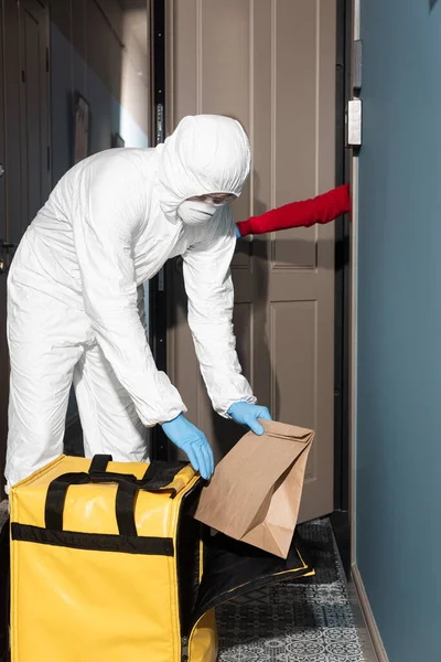 Uomo di consegna in tuta hazmat e maschera medica prendendo pacchetto dalla borsa termica vicino alla donna porta di apertura — Foto stock