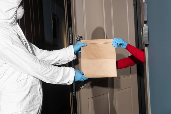 Side view of man in hazmat suit and medical mask giving package to woman in latex gloves near open door — Stock Photo