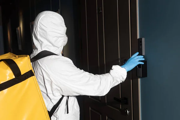 Боковой вид курьера в защитном костюме с терморюкзаком, звонящим в дверной звонок — стоковое фото