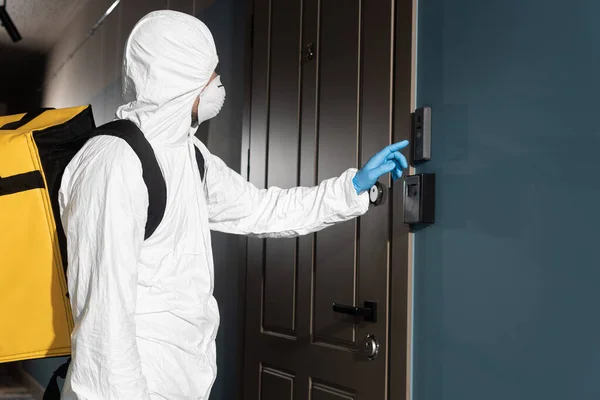 Vista lateral do correio em terno hazmat, máscara médica e luva de látex tocando na campainha — Fotografia de Stock