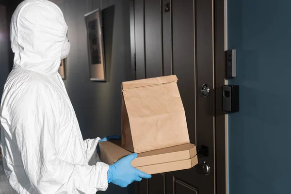 Vista lateral del mensajero en traje de materiales peligrosos y paquete de retención de máscara médica y cajas de pizza cerca de la puerta - foto de stock