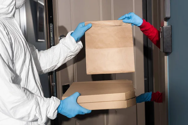 Vista recortada de mensajero en traje de materiales peligrosos y máscara médica dando cajas de pizza y paquete a la mujer en guantes de látex cerca de la puerta abierta - foto de stock