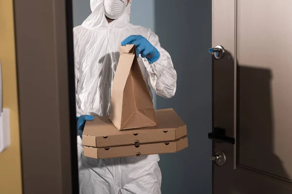 Vista recortada del repartidor con traje de materiales peligrosos y guantes de látex sosteniendo el paquete y cajas de pizza cerca de la puerta abierta - foto de stock