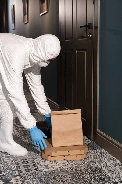 Vista lateral del repartidor en traje de materiales peligrosos y máscara médica poniendo paquete y cajas de pizza en el piso cerca de la puerta en el porche - foto de stock