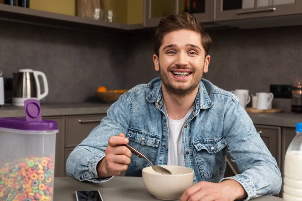 Selective focus of cheerful man eating cereals near smartphone on table — Stock Photo