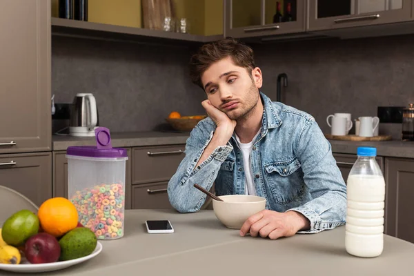 Selective focus of sad man looking away near cereals and smartphone on table in kitchen — Stock Photo