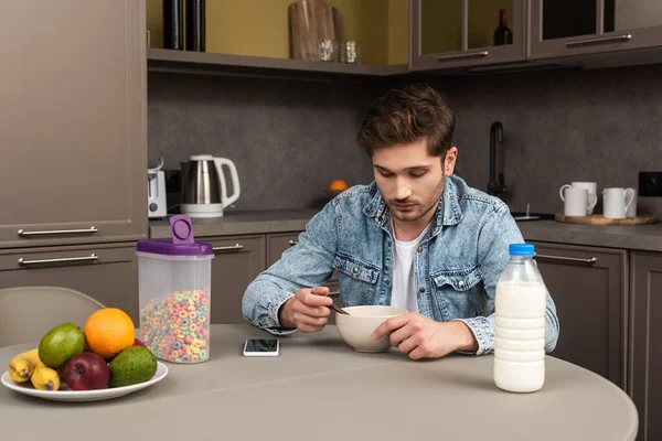 Man eating cereals near fresh fruits and smartphone on table in kitchen — Stock Photo