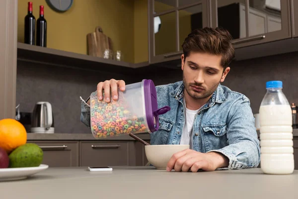 Selektiver Fokus des Mannes, der Getreide in einer Schüssel neben einer Flasche Milch und Obst auf den Küchentisch schüttet — Stockfoto