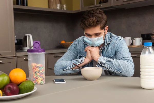 Focus selettivo dell'uomo in maschera medica seduto vicino a cereali, bottiglia di latte e frutta in cucina — Foto stock