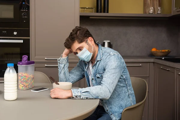 Vue latérale de l'homme bouleversé dans le masque médical assis près des céréales et smartphone sur la table dans la cuisine — Photo de stock