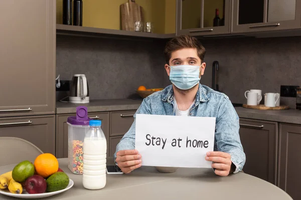 Man in medical mask holding card with stay at home lettering near cereals and fruits on table in kitchen — Stock Photo