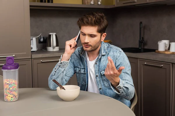 Handsome man talking on smartphone near cereals on kitchen table — Stock Photo