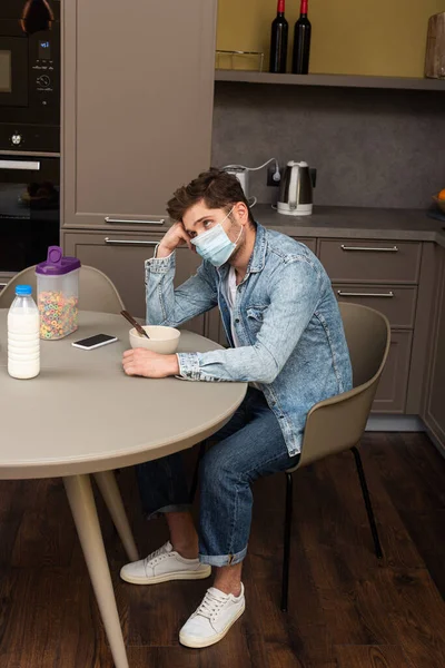 Vista lateral do homem chateado em máscara médica olhando para longe perto de cereais e smartphone na mesa da cozinha — Fotografia de Stock