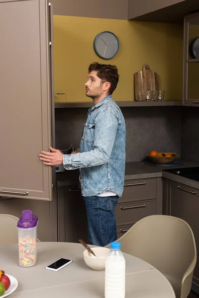 Вид сбоку на красивого мужчину, открывающего холодильник рядом с зерновыми и смартфоном на столе — стоковое фото