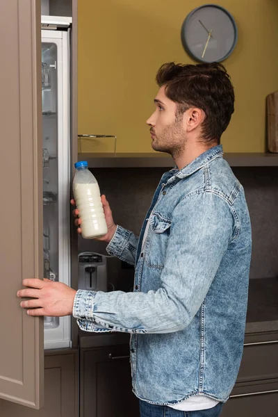 Vue latérale du bel homme tenant une bouteille de lait près d'un réfrigérateur ouvert dans la cuisine — Photo de stock