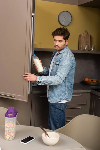 Vista lateral de homem bonito segurando garrafa de leite perto de geladeira e cereais na mesa na cozinha — Fotografia de Stock