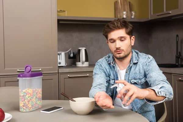 Bell'uomo che utilizza disinfettante per le mani vicino a cereali e smartphone sul tavolo della cucina — Foto stock