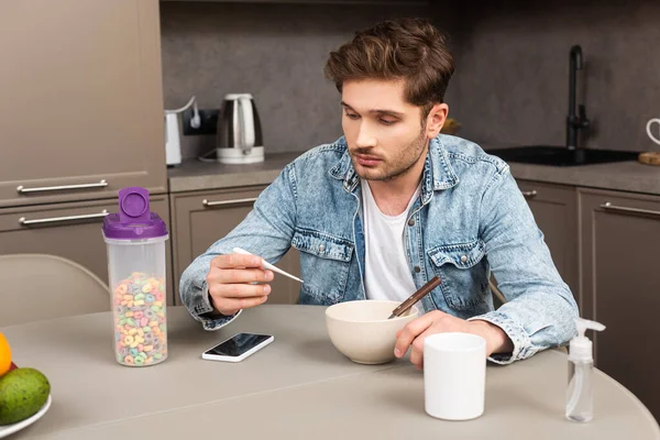 Hombre guapo sosteniendo termómetro cerca de teléfono inteligente y cereales en la mesa de la cocina - foto de stock