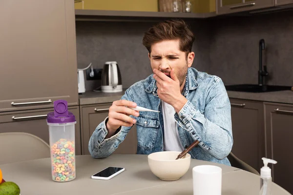 Hombre bostezando mientras sostiene el termómetro cerca de los cereales y el teléfono inteligente en la mesa - foto de stock