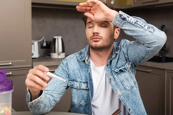 Selective focus of sick man with hand near forehead holding thermometer in kitchen — Stock Photo