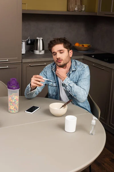 Больной мужчина трогает шею и держит термометр рядом со смартфоном и злаками на столе на кухне — стоковое фото