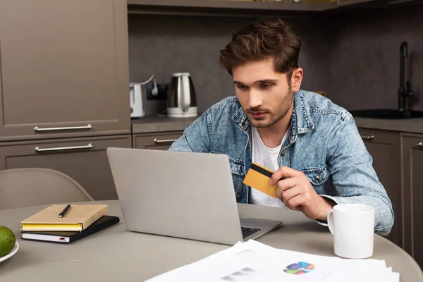 Selective focus of handsome man holding credit card and using laptop on kitchen table — Stock Photo