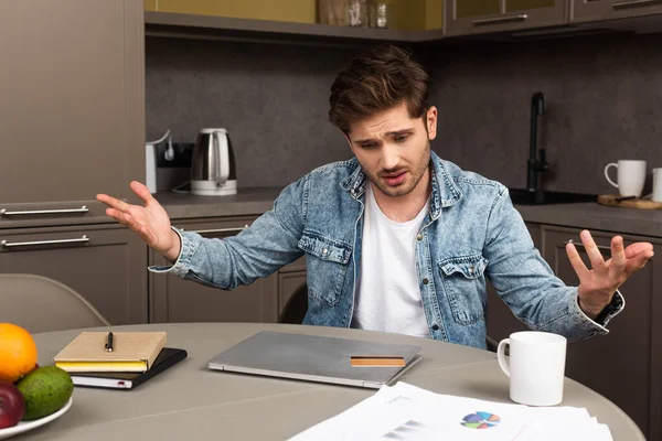 Вибірковий фокус хвилюючого чоловіка, який дивиться на кредитну картку на ноутбуці біля паперів на кухонному столі — стокове фото