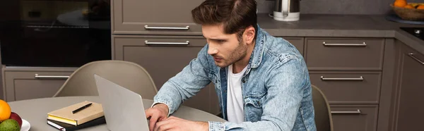 Panoramic crop of freelancer using laptop near book and notebook on kitchen table — Stock Photo