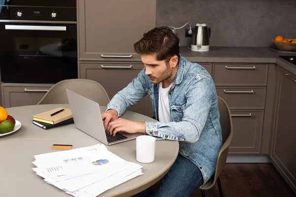 Красивий чоловік використовує ноутбук під час роботи біля паперу та кредитної картки на столі на кухні — стокове фото