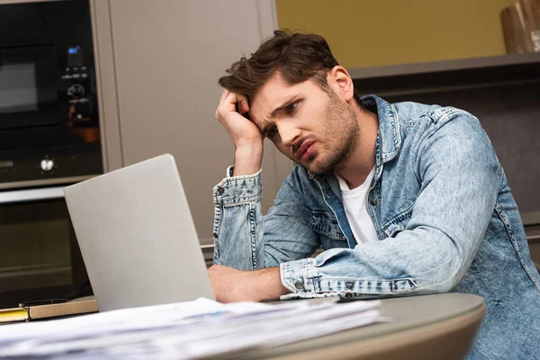 Selective focus of upset man looking at laptop near papers on kitchen table — Stock Photo