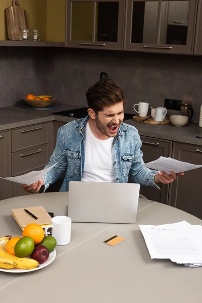 Teleworker irritado segurando documentos perto de laptop e cartão de crédito na mesa da cozinha — Fotografia de Stock