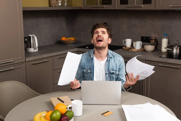 Irritierter Mann mit Papieren in der Nähe von Kreditkarte und Laptop auf Tisch in Küche — Stockfoto