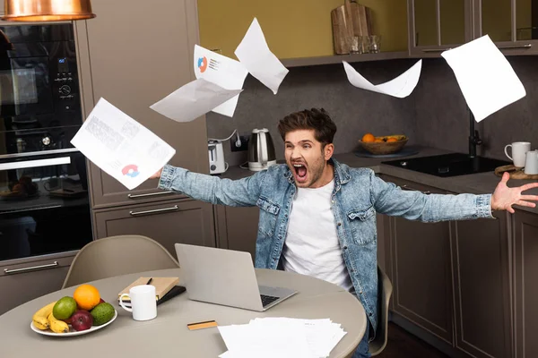Кричащий фрилансер бросает документы во время работы с ноутбуком на кухонном столе — стоковое фото