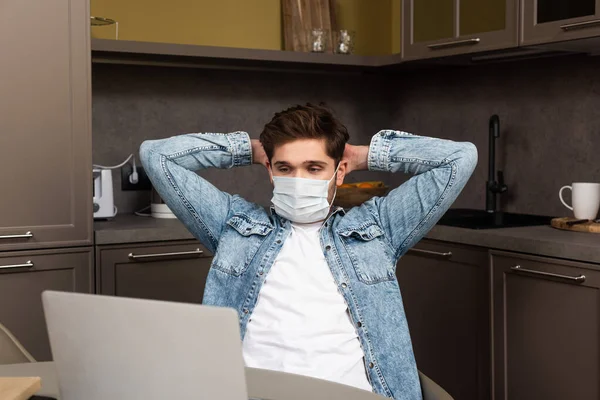 Selective focus of freelancer in medical mask looking at laptop on kitchen table — Stock Photo