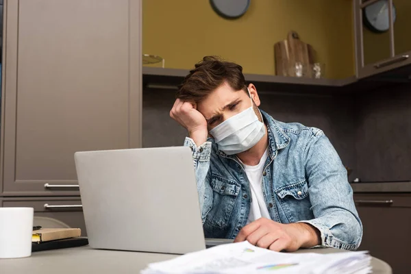 Foco seletivo de freelancer triste em máscara médica sentada à mesa com papéis e laptop — Fotografia de Stock